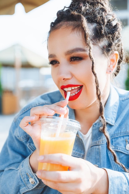 ドレッドヘアと赤い口紅を持つ女性、屋外カフェでオレンジ ジュースを飲んで楽しんでいるかわいいスタイリッシュなファッショナブルな女の子の屋外のクローズ アップの肖像画