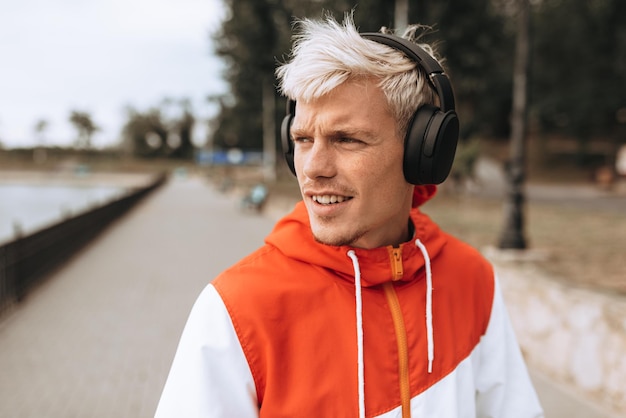 Outdoor closeup portrait of handsome young man enjoying weather in city park waiting for his girlfriend while listening to his favorite music from wireless headphones in the park