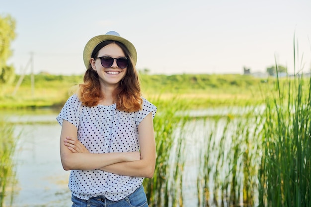Outdoor country portrait of a teenage girl in a hat sunglasses near a pond in the reeds, summer sunset natural background, copy space