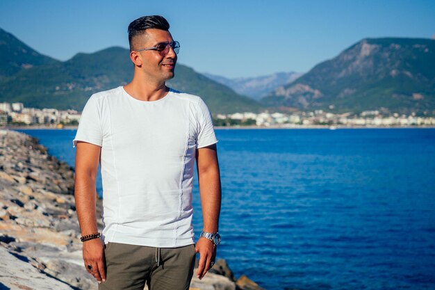 Outdoor fashion portrait of handsome man posing at amazing tropical Turkey beach beautiful view on blue sky and mountains and the hills wearing casual classic white shirt and sunglasses