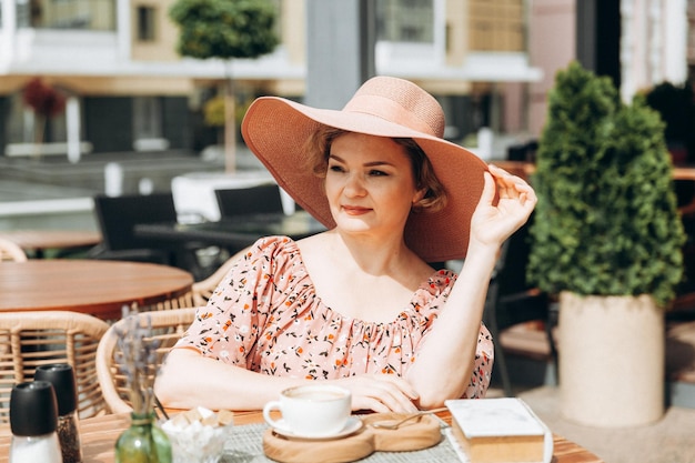 Outdoor fashion portrait of a stunning woman sitting in a cafe I drink coffee and read an old book a woman in a dress and a hat