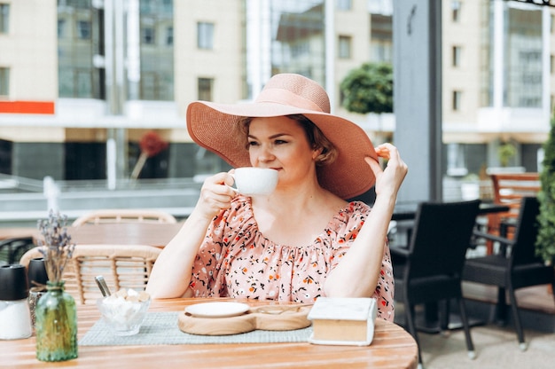 Outdoor fashion portrait of a stunning woman sitting in a cafe I drink coffee and read an old book a woman in a dress and a hat