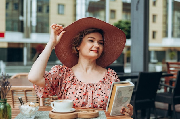 Outdoor fashion portrait of a stunning woman sitting in a cafe I drink coffee and read an old book a woman in a dress and a hat