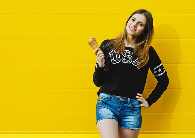 Outdoor fashion portrait of young hipster girl with ice-cream on yellow wall background