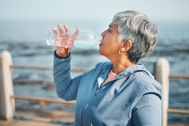 Outdoor fitness or senior woman drinking water for hydration wellness and thirsty on beach run training or exercise Bottle runner or elderly person refresh with cold liquid on retirement workout