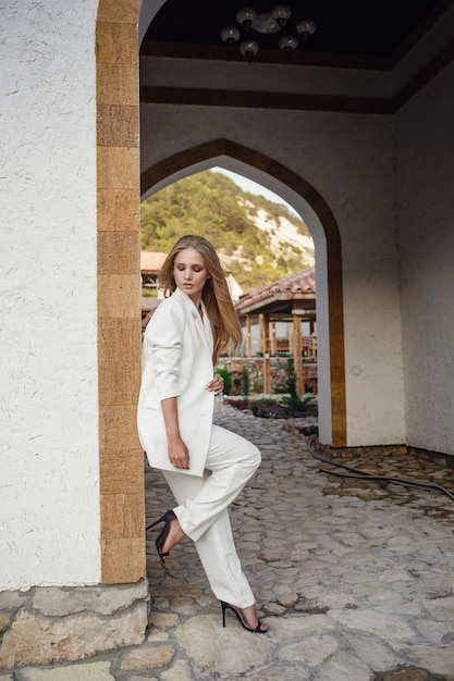 Outdoor full body fashion portrait of fashionable woman in white suit posing in street of east city Beautiful stylish shot of a tanned blonde for a fashion magazine