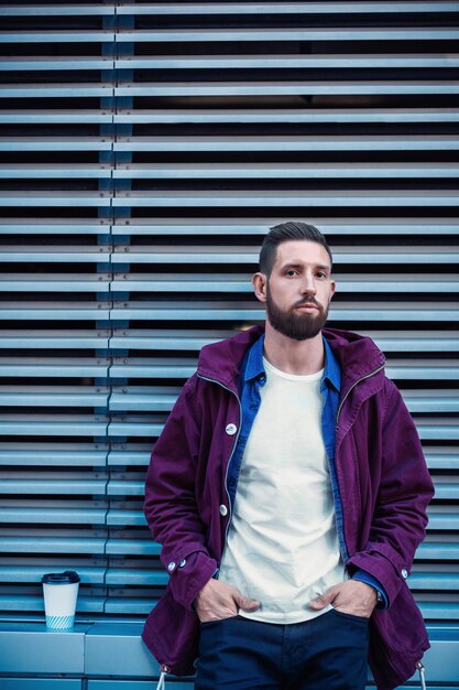 Outdoor herfst- of winterportret van een knappe hipsterman met baard, wit t-shirt, blauw shirt en bruin jasje met een kop warme koffie in de hand.