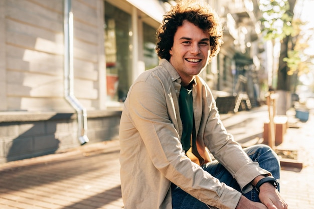 Outdoor image of happy smiling man with curly hair posing for social advertisement sitting on the bench outdoors in the city street Excited cheerful student male resting outside