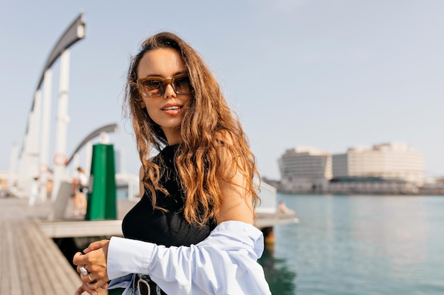 Photo outdoor lifestyle of cheerful young caucasian woman in sunglasses on street posing at camera tanned brunette model smiling gently real emotions concept