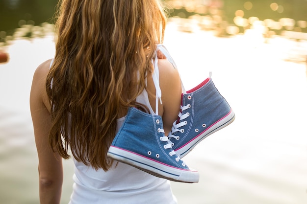 Photo outdoor lifestyle closeup view of of tied pair of jeans sneakers hanging on a finger of a beautiful young woman over the shoulder