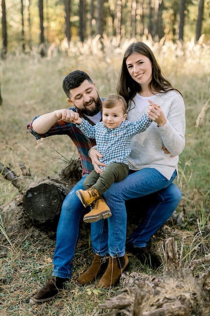 Outdoor lifestyle portrait of happy Caucasian family, father, mother and little baby boy, wearing stylish casual clothes, sitting on a log in pine autumn forest and smiling at camera