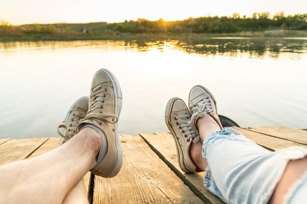 Outdoor lifestyle view of a couple wearing sneakers sitting on a wooden pier at the water on a sunset