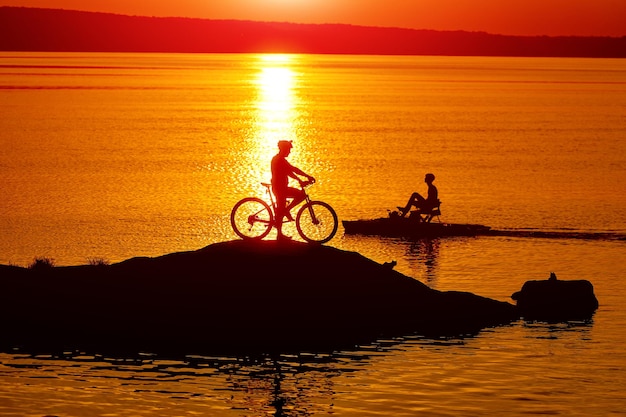 休んでいる男の屋外オレンジ色の日光。湖に座っている男の夕日のシルエット。