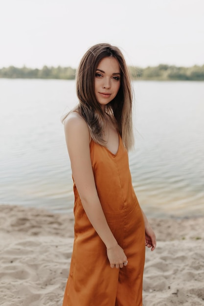 Foto all'aperto di incredibile ragazza alla moda con la pelle pallida timida che ride e guarda la fotocamera ritratto di giovane donna bruna estatica in prendisole arancione sorridente sullo sfondo del lago