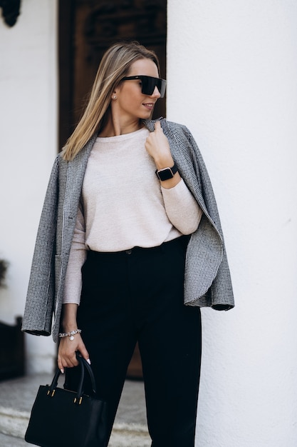 Outdoor photo of blonde lady posing on architecture background in autumn day. Fashion street style portrait. wearing dark casual trousers and creamy sweater and sunglasses. Fashion concept.