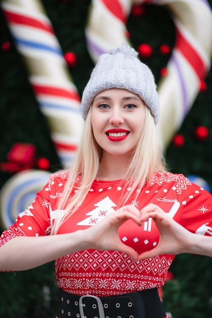 Outdoor photo of happy blonde girl in winter hat Shows his heart with his hands Festive Christmas