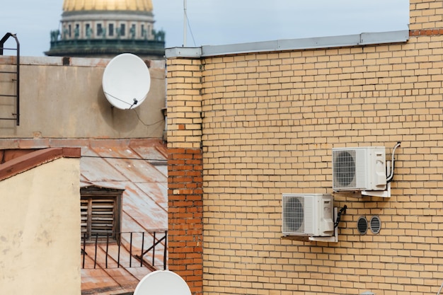 Outdoor photo of two white air conditioners mounted on yellow brick wall and two sattelite dishes