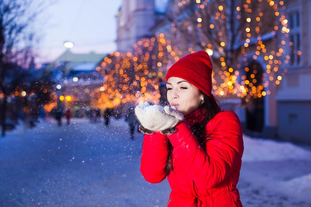 Outdoor photo of young beautiful girl blowing away snow from her warm white mittens, posing in street with Christmas lights on trees on background