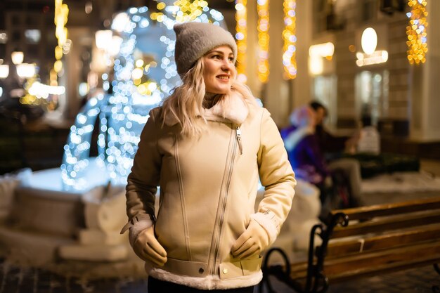 Outdoor photo of young beautiful happy smiling girl holding sparklers, posing in street. Festive Christmas fair on background. Model wearing stylish winter coat, knitted beanie hat, scarf.