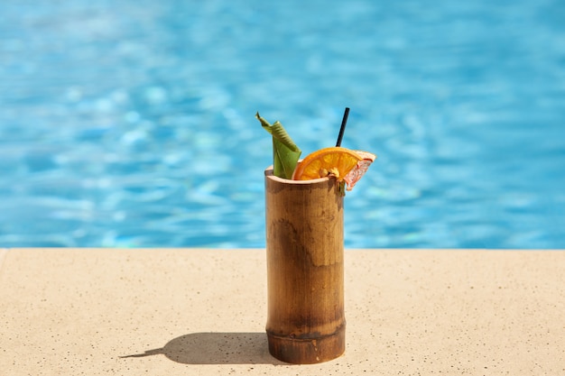 Immagine all'aperto della tazza di legno con il cocktail vicino alla piscina, verde e pezzo di arancia all'interno del cocktail, paglia nera da bere, liberarsi della sete, bevanda fredda nella stagione calda.