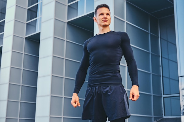 Outdoor portrait of attractive, athletic male dressed in a black sportswear over modern building background.