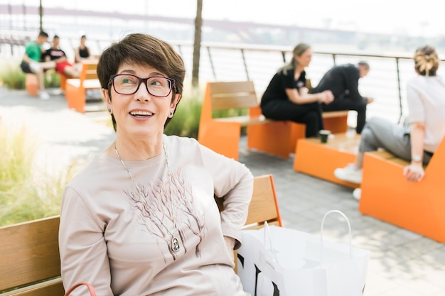 Foto ritratto all'aperto di una bella e elegante donna di mezza età di 55 a 60 anni con una maglietta a maniche lunghe seduta su una panchina in strada spazio di copia e posto vuoto per la pubblicità di testo