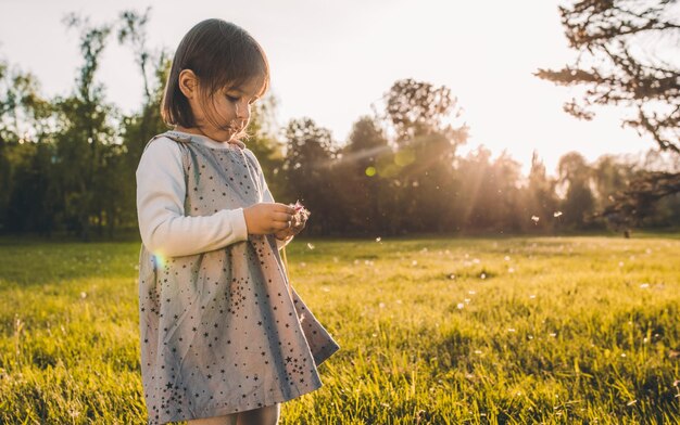 Outdoor portrait of beautiful little girl child playing with dandelion on meadow on sunset light Cute child enjoy and explore nature in the park Happy emotion Happy Mother's Day Childhood