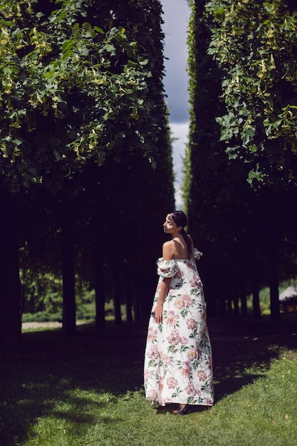 꽃이 있는 드레스를 입은 아름다운 호화 브루네트 여성의 야외 초상화는 손질된 나무가 있는 공원에 서 있다