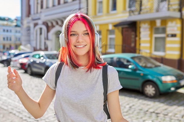 Outdoor portrait of beautiful trending teenage girl listening music with wireless headphones Happy smiling female with trendy colored hairstyle on street of sunny summer city