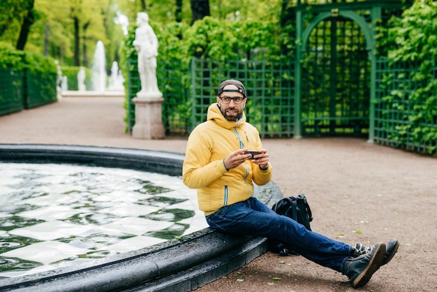 Outdoor portrait of bushy man wearing cap jacket jeans and eyeglasses having rest near fountain holding his modern device in hands being happy to notice his friend Relaxed hipster man in open air