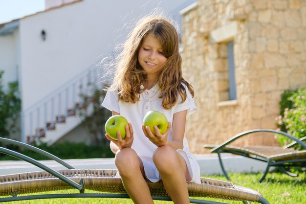 Outdoor portrait of a child girl with two green apples sitting in a garden chair