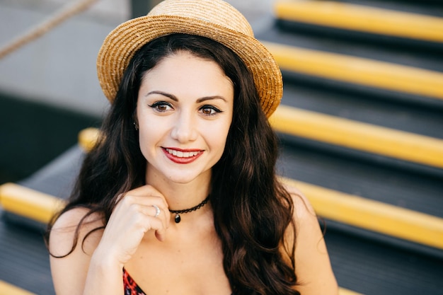 Ritratto all'aperto di donna alla moda con capelli scuri e folti che si trucca e labbra rosse dipinte che indossa un cappello di paglia estivo e una collana che guarda in lontananza con un sorriso gentile seduto sulle scale