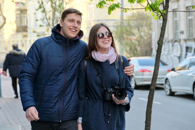 Outdoor portrait of happy couple in love walking along street of winter spring city hugging, woman with camera