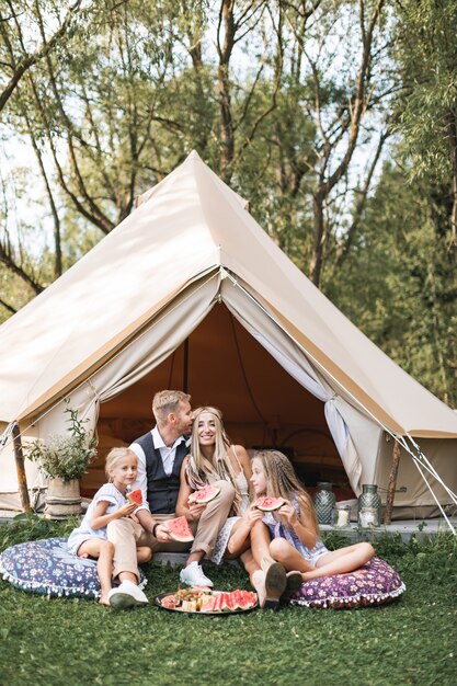 Outdoor portrait of happy family, father, mother and two little daughters, sitting on the grass and enjoying watermelon at the picnic near camp tent