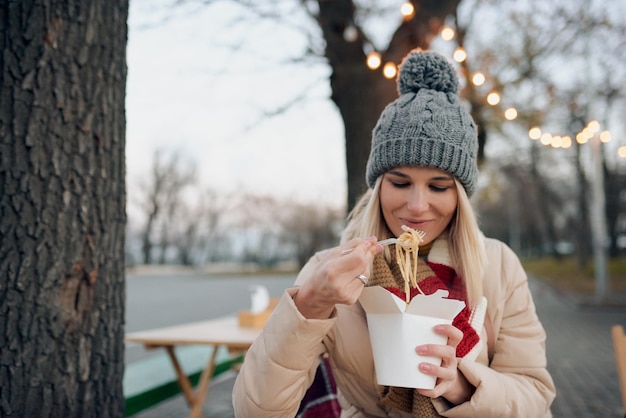 Outdoor portrait of happy pretty woman eating asian wok fast food from takeaway white paper box at the city street Student female or worker have a lunch break delicious wok noodles in cold wheather