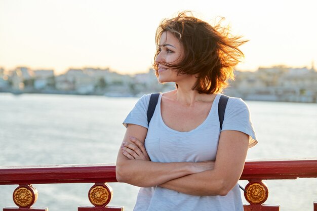 Outdoor portrait of mature beautiful happy woman with arms crossed, female walking with backpack on bridge on summer day, golden hour. Woman looks away, copy space