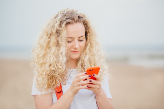 Outdoor portrait of pleasant looking curly female uses modern cell phone for online communication.