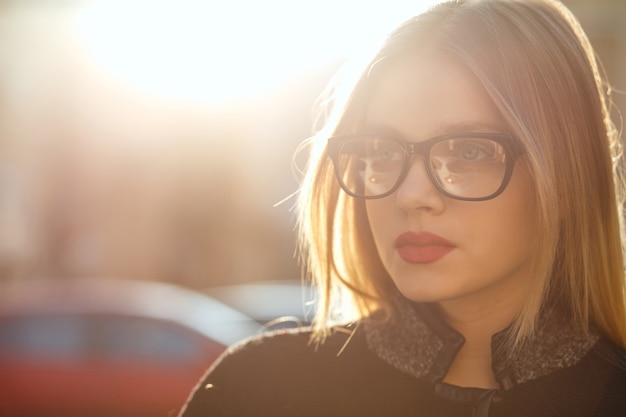 Outdoor portrait of seductive blonde woman wearing glasses and coat, posing at the old sunny street. Space for text