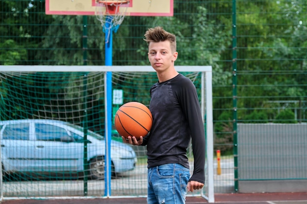 Outdoor portrait of teenage boy with ball on street basketball court, trendy male 16, 17 years old looking at the camera. Active healthy lifestyle, hobbies and leisure, teenagers concept