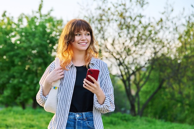 Outdoor portrait of trendy young redhaired girl with smartphone in hand green trees sky background smiling attractive teenage female student youth beauty fashion lifestyle leisure concept