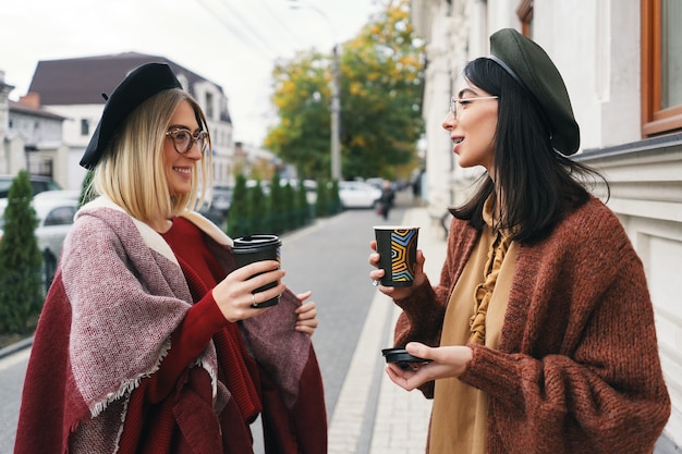 Outdoor portrait of two female friends talking. Girls in casual warm outfits and glasses having city walk in cold season and having coffee at urban street. City lifestyle, friendship concept.