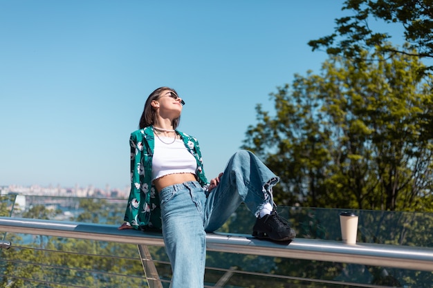 Outdoor portrait of woman in casual green shirt and jeans at sunny day on bridge with city amazing view in morning