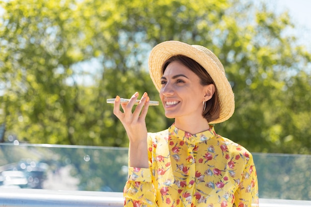 Outdoor portrait of woman in yellow summer dress and hat recording audio voice message on phone