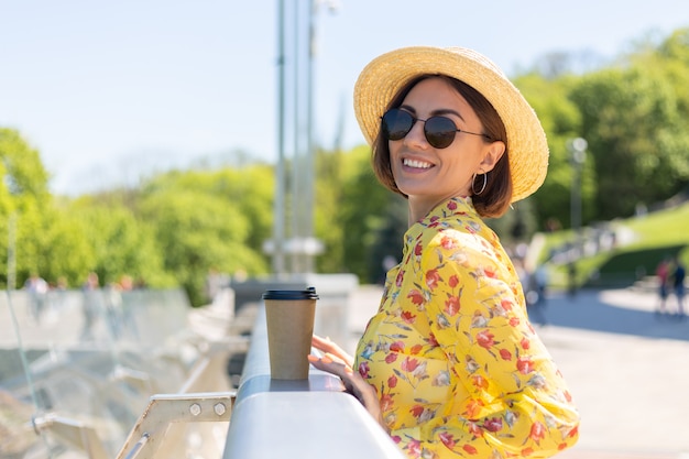 Outdoor portrait of woman in yellow summer dress and hat with cup of coffee enjoying sun, stands on bridge with city amazing view