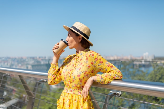Outdoor portrait of woman in yellow summer dress and hat with cup of coffee enjoying sun, stands on bridge with city amazing view