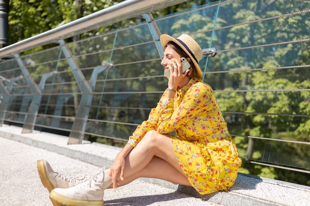 Outdoor ritratto di donna in abito estivo giallo seduto sul ponte effettua una chiamata, parlando al cellulare