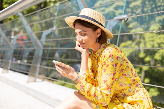 Outdoor portrait of woman in yellow summer dress sitting on bridge with mobile  phone looking on screen with a smile