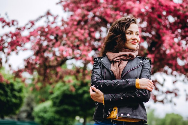 花木の近くポーズ若い美しいファッショナブルな女性の屋外のポートレート。女性の美しさとファッション。シティライフスタイル。黄色のカーディガンピンクの花盛りの木の女の子。