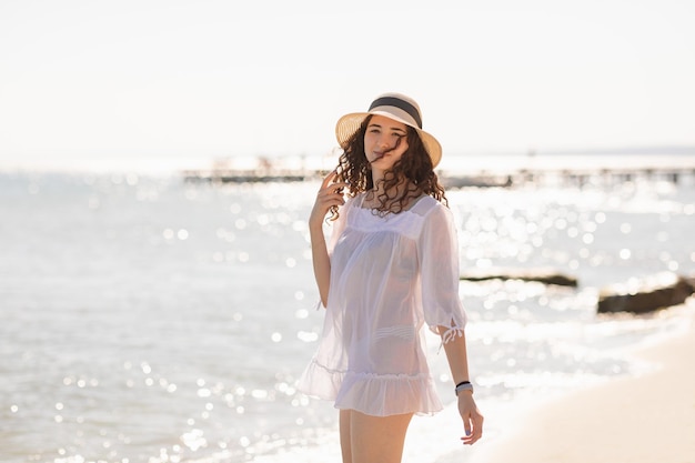 Outdoor portrait of young brunette teenager girl with curly hair and straw hat at sunny day staying on the beach in white beachwear