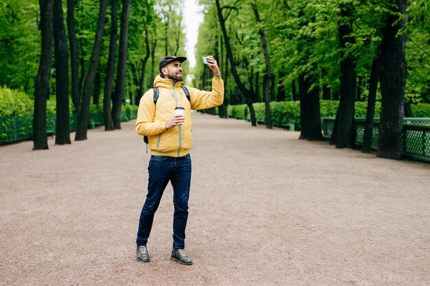 Ritratto all'aperto di giovane bel turista vestito con abiti eleganti che tengono zaino e caffè da asporto in posa nella fotocamera del suo smartphone facendo selfie isolato su sfondo verde natura